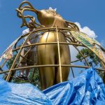 A person working on a large golden sculpture surrounded by blue tarps and greenery under a clear sky.