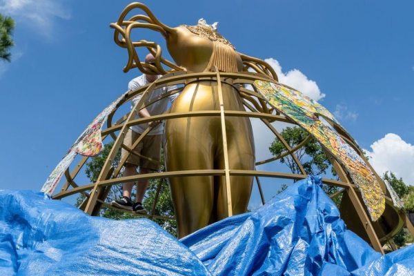 A person working on a large golden sculpture surrounded by blue tarps and greenery under a clear sky.