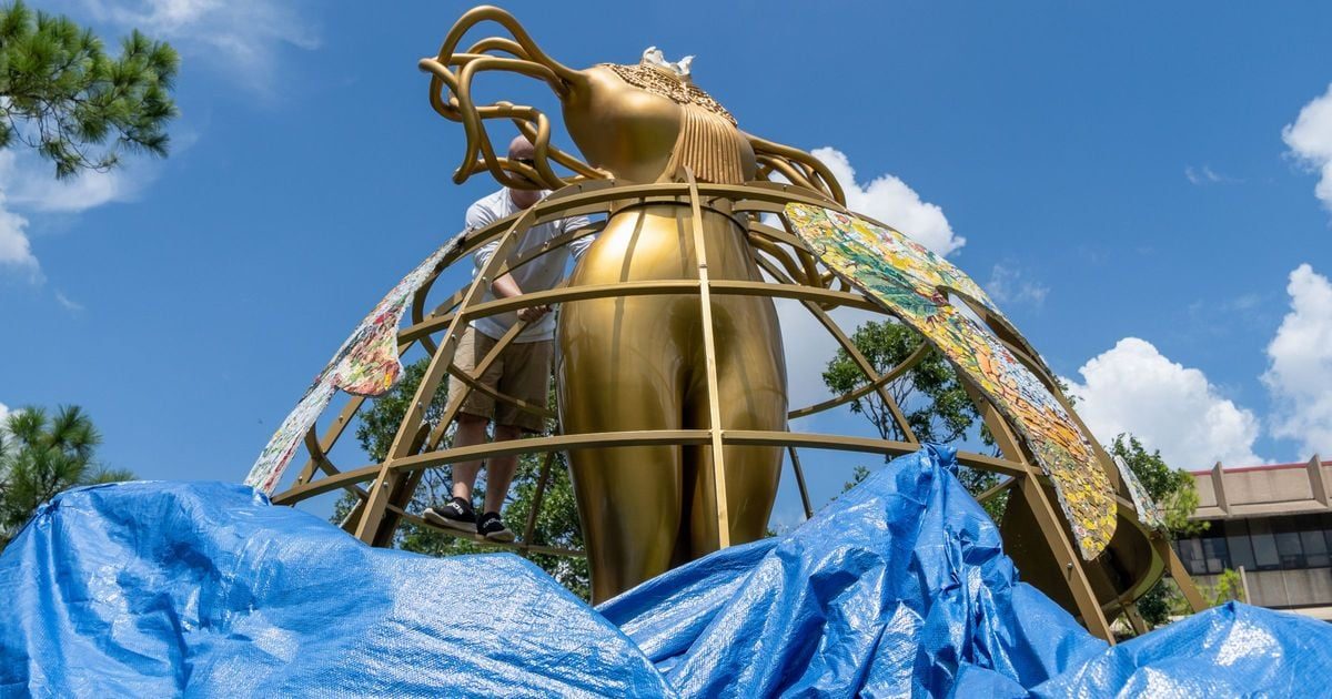 A person working on a large golden sculpture surrounded by blue tarps and greenery under a clear sky.